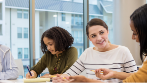 Woman taking notes while other women are in a discussion during a meeting
